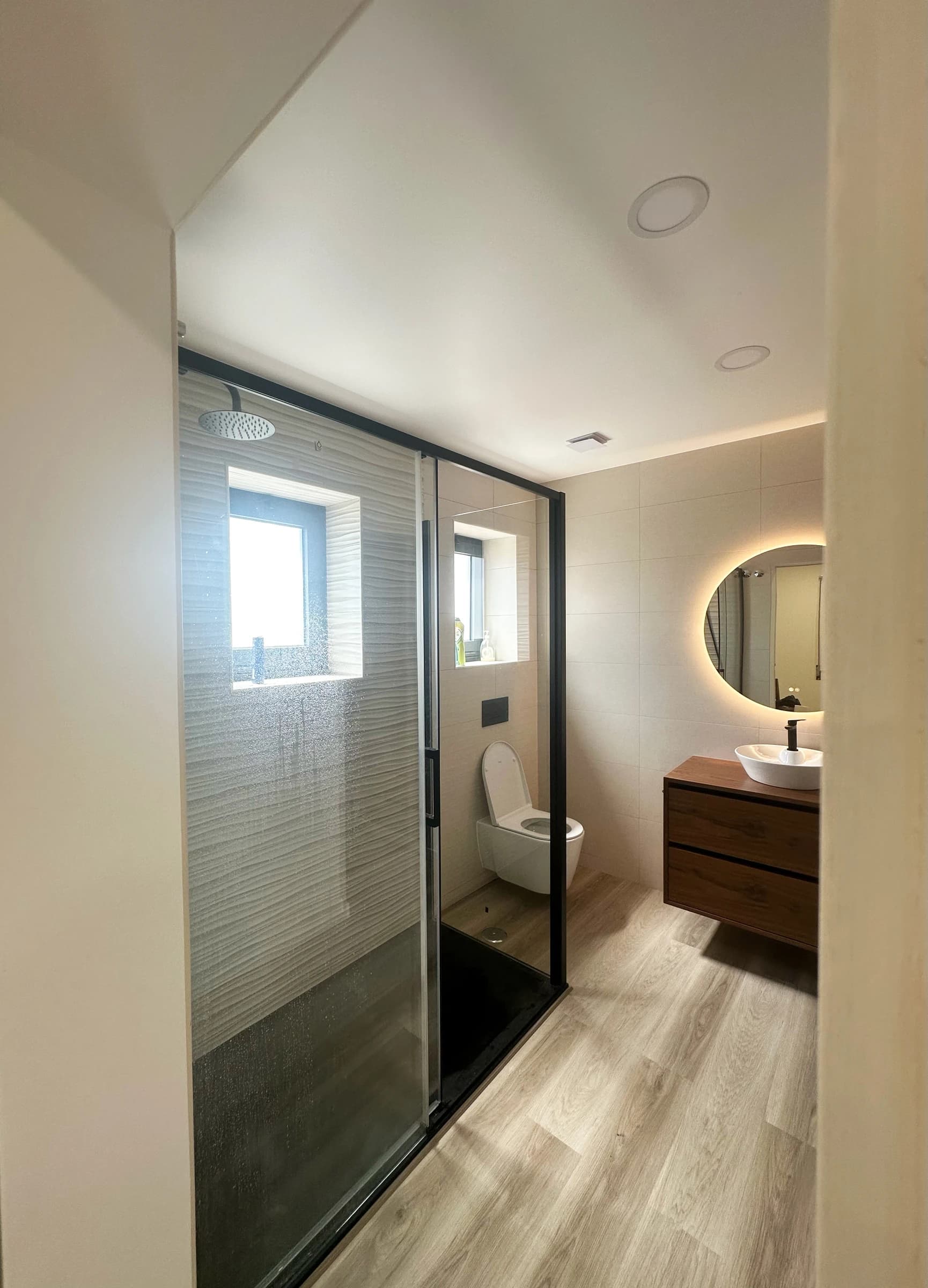 Casa de banho de suite em Estarreja, Aveiro, com duche de base preta, espelho redondo LED e móvel suspenso em madeira natural — remodelação VPObras